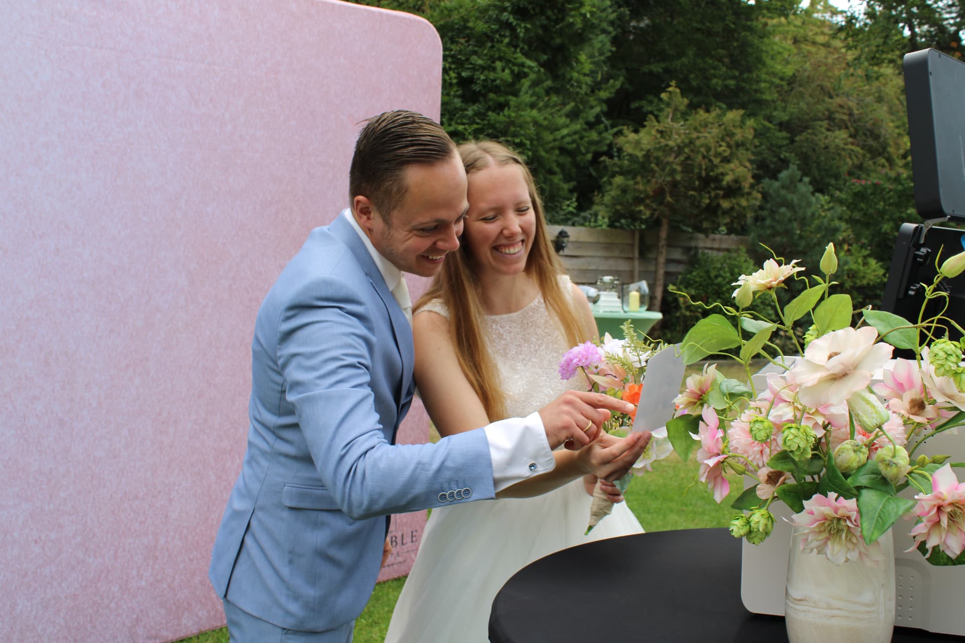 Bruidspaar bekijkt samen lachend hun foto bij de photobooth met roze achtergrond
